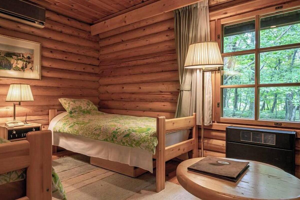 Quarto de cabana de tronco com duas camas, uma mesa, uma luminária, um quadro na parede e uma janela com vista para árvores.