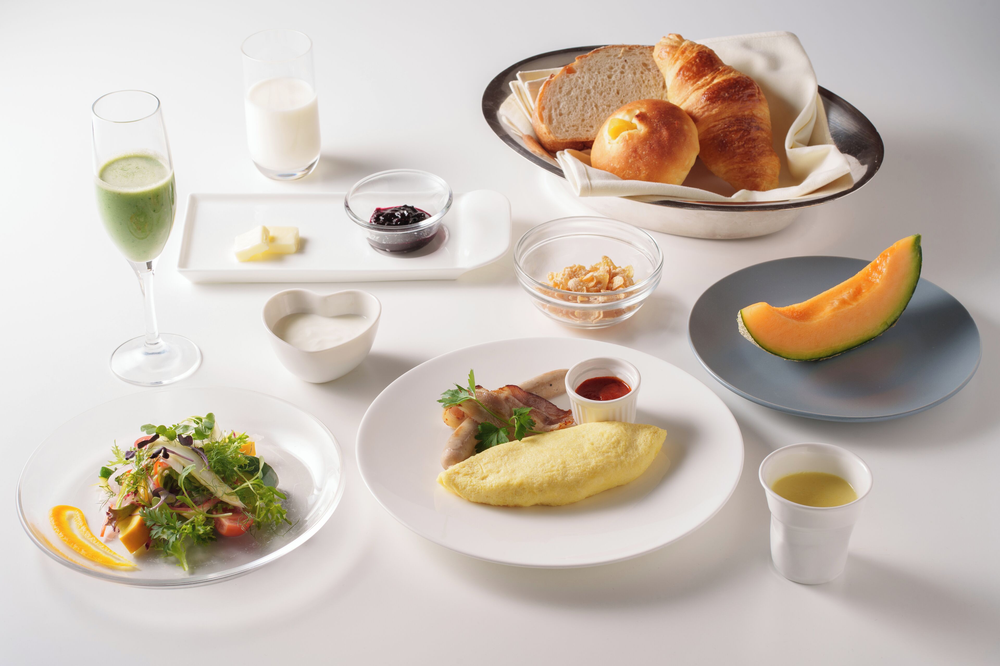 A variety of breakfast items including croissants, a croissant basket, a plate with an omelet, a plate with a melon slice, a glass of green juice, a glass of milk, and a bowl of salad.
