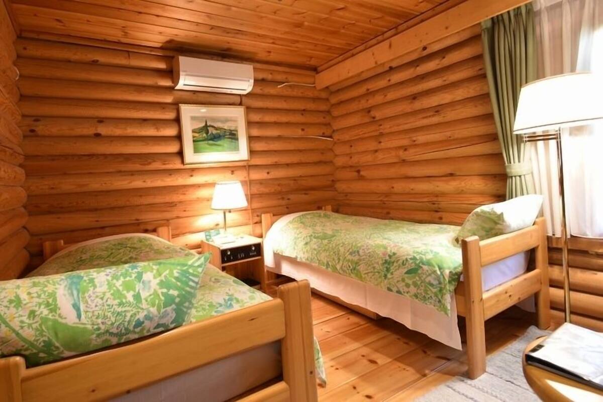 Quarto de cabana de madeira com duas camas de madeira, uma mesa de cabeceira com abajur, um aparelho de ar condicionado e uma moldura com uma imagem na parede.