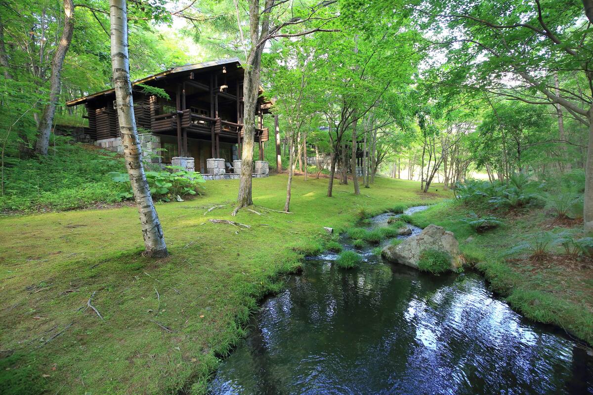 A rustic cabin with a wooden deck and a small stream in a forested area.