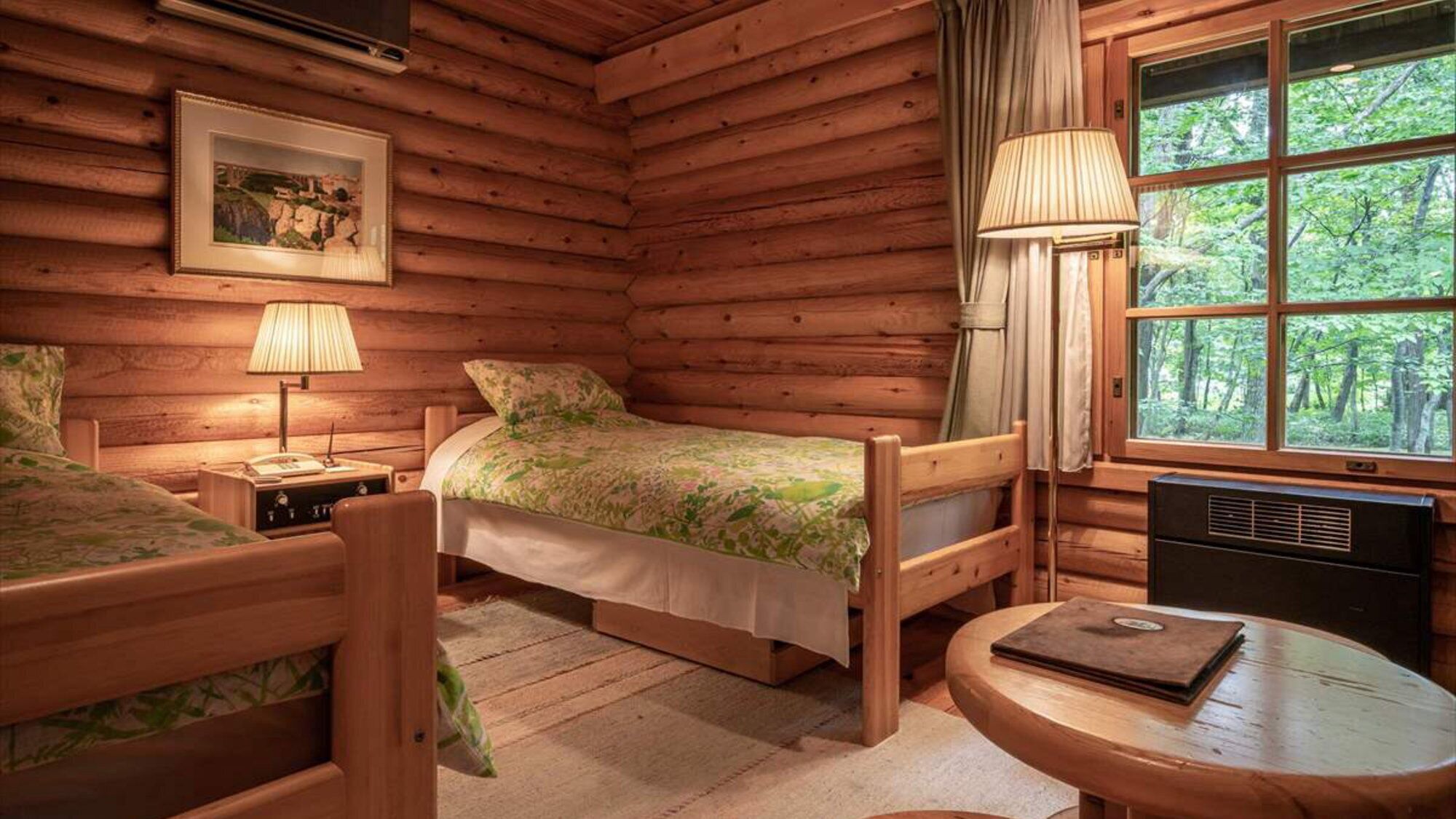 A log cabin room with a bed, a chair, a table, a lamp, and a window with a view of trees.
