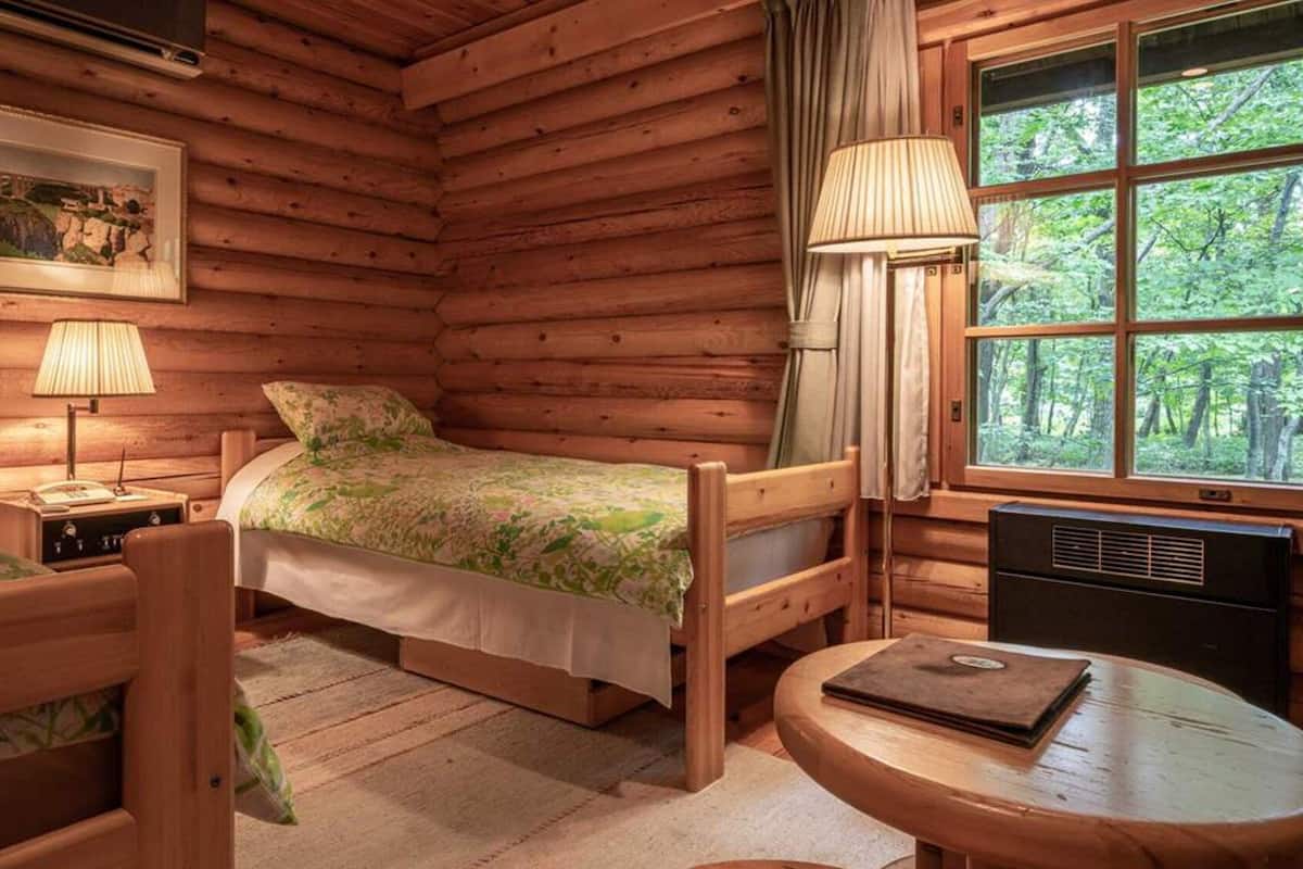 Quarto de cabana de tronco com cama, cadeira, mesa, abajur e janela com vista para árvores.