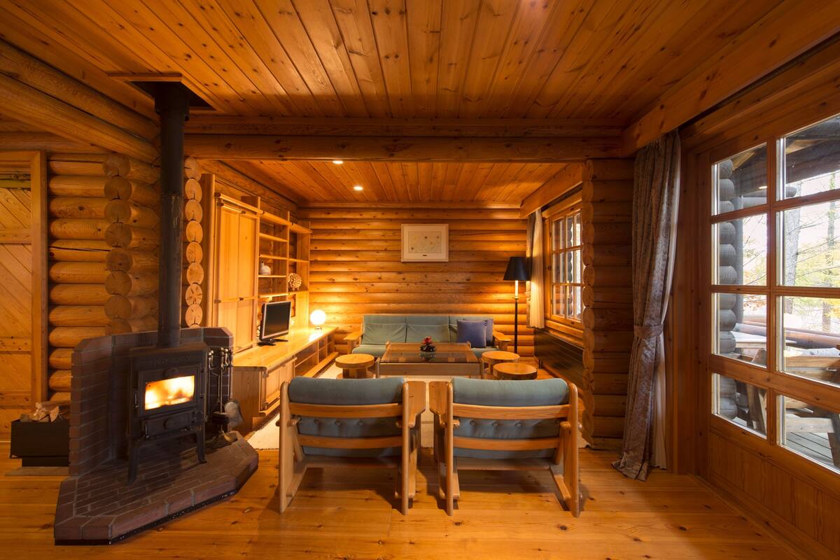Interior de uma cabana de madeira com lareira, móveis rústicos e janelas amplas.