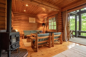 Interior acogedor de una cabaña de madera con estufa de leña, un sofá, una mesita y una ventana con cortinas.