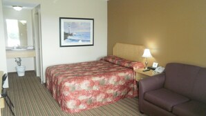 A hotel room with a bed, a sofa, a desk with a lamp, a framed picture on the wall, and a bathroom with a sink and mirror. - Bayview inn (Orillia)