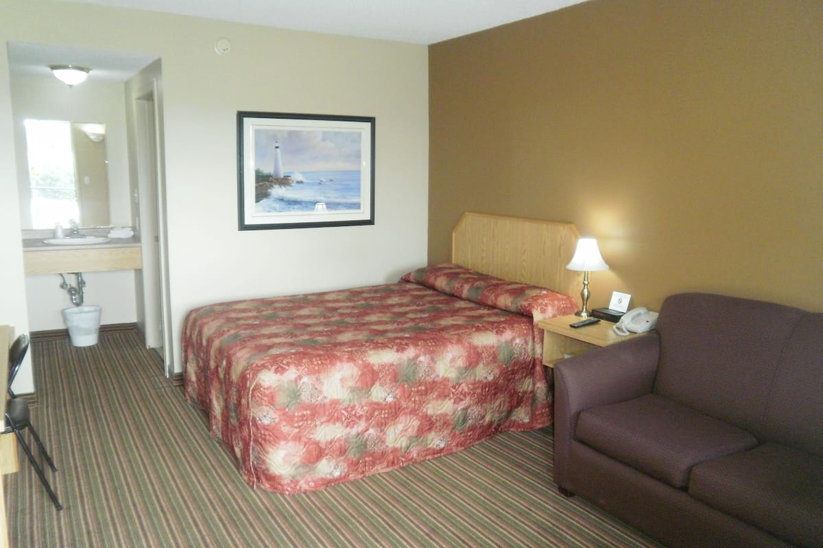 Habitación de hotel con cama, sofá, escritorio con lámpara, un cuadro en la pared y baño con lavabo y espejo.