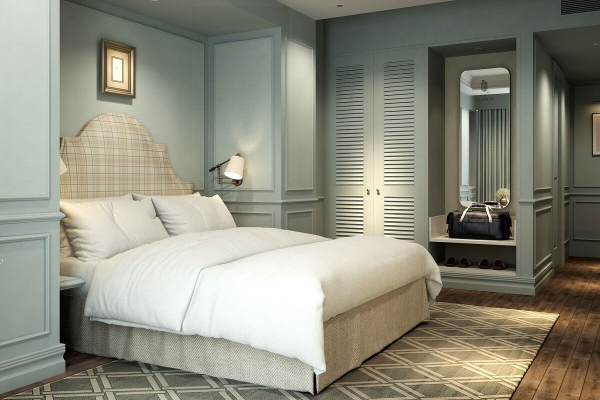 A bedroom with a bed, a nightstand, a wall-mounted lamp, a wardrobe with louvered doors, a mirror, and a bench with shoes.