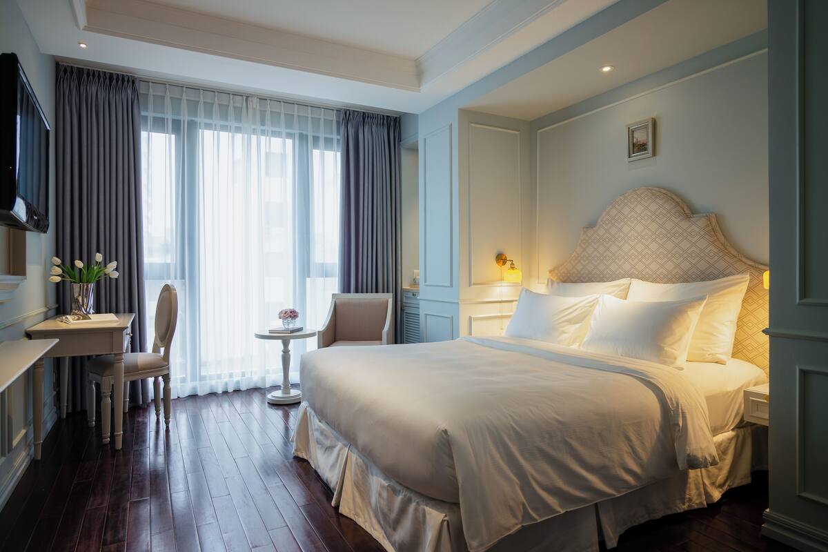 A hotel room with a large bed, a desk with a chair, a small table with a vase, and a television on the wall.