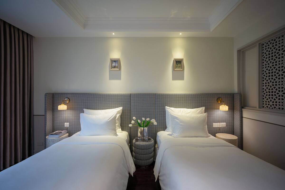 A hotel room with two beds, a small round table with a vase of flowers, and wall-mounted light fixtures.