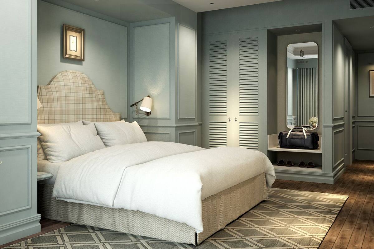 A bedroom with a bed, a nightstand, a wall-mounted lamp, a wardrobe with louvered doors, a mirror, and a bench with shoes.