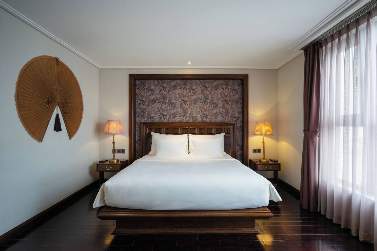 A large bed with a wooden headboard, two bedside tables with lamps, a wall fan, and a window with curtains.