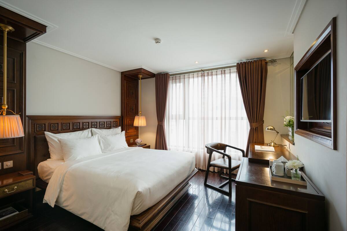 A hotel room with a large bed, a desk with a chair, a lamp, and a window with curtains.