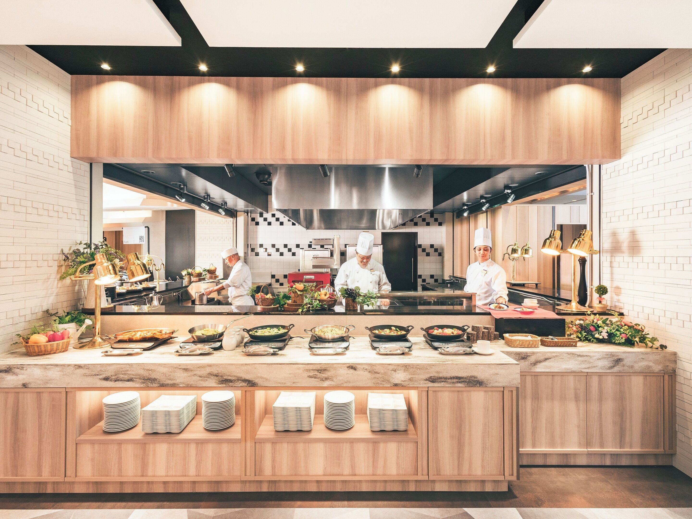 A modern kitchen with chefs preparing food, a wooden counter with dishes, and a brick wall.