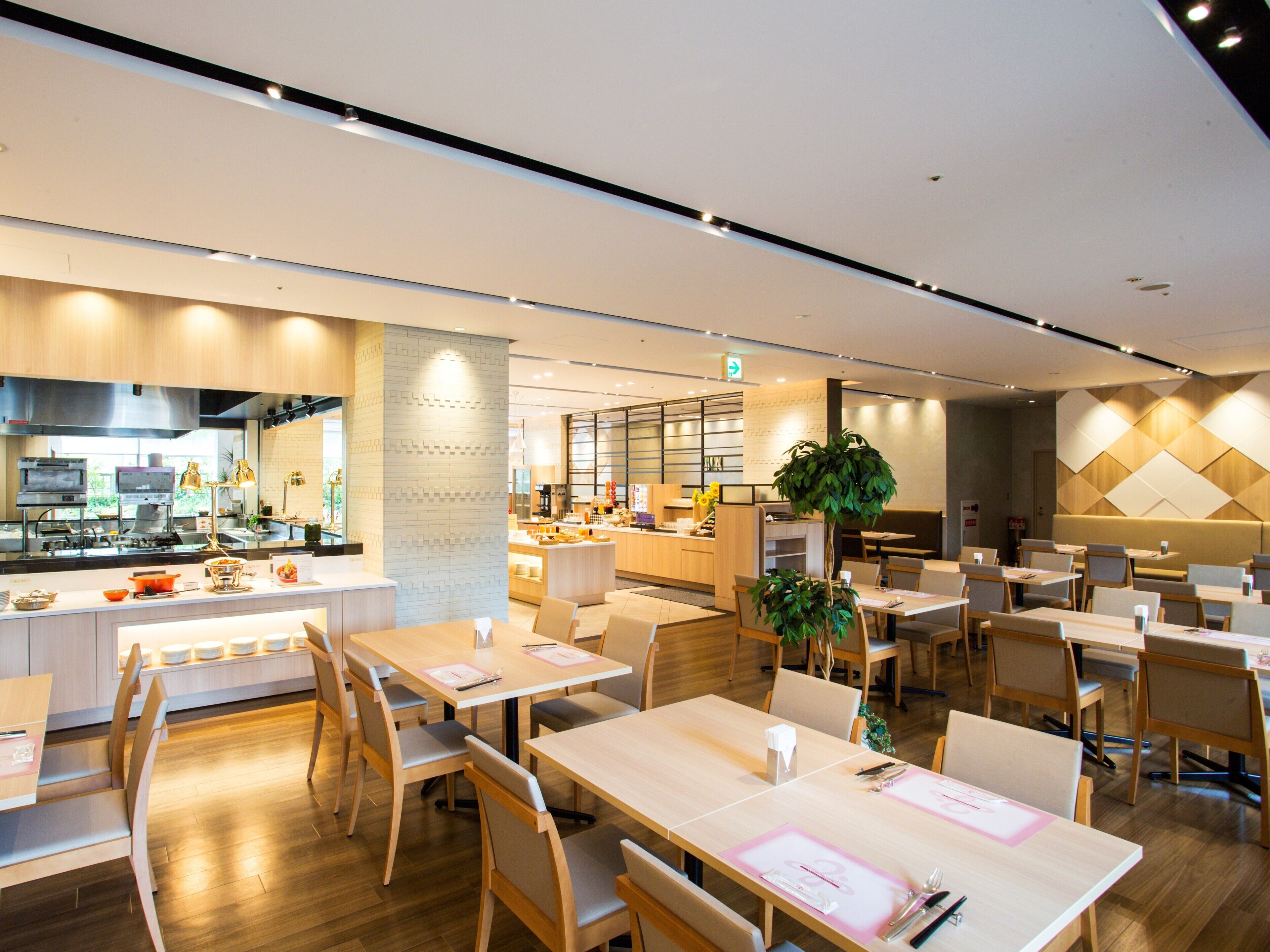 A modern restaurant with wooden tables and chairs, a kitchen area, and a seating area with a large plant.