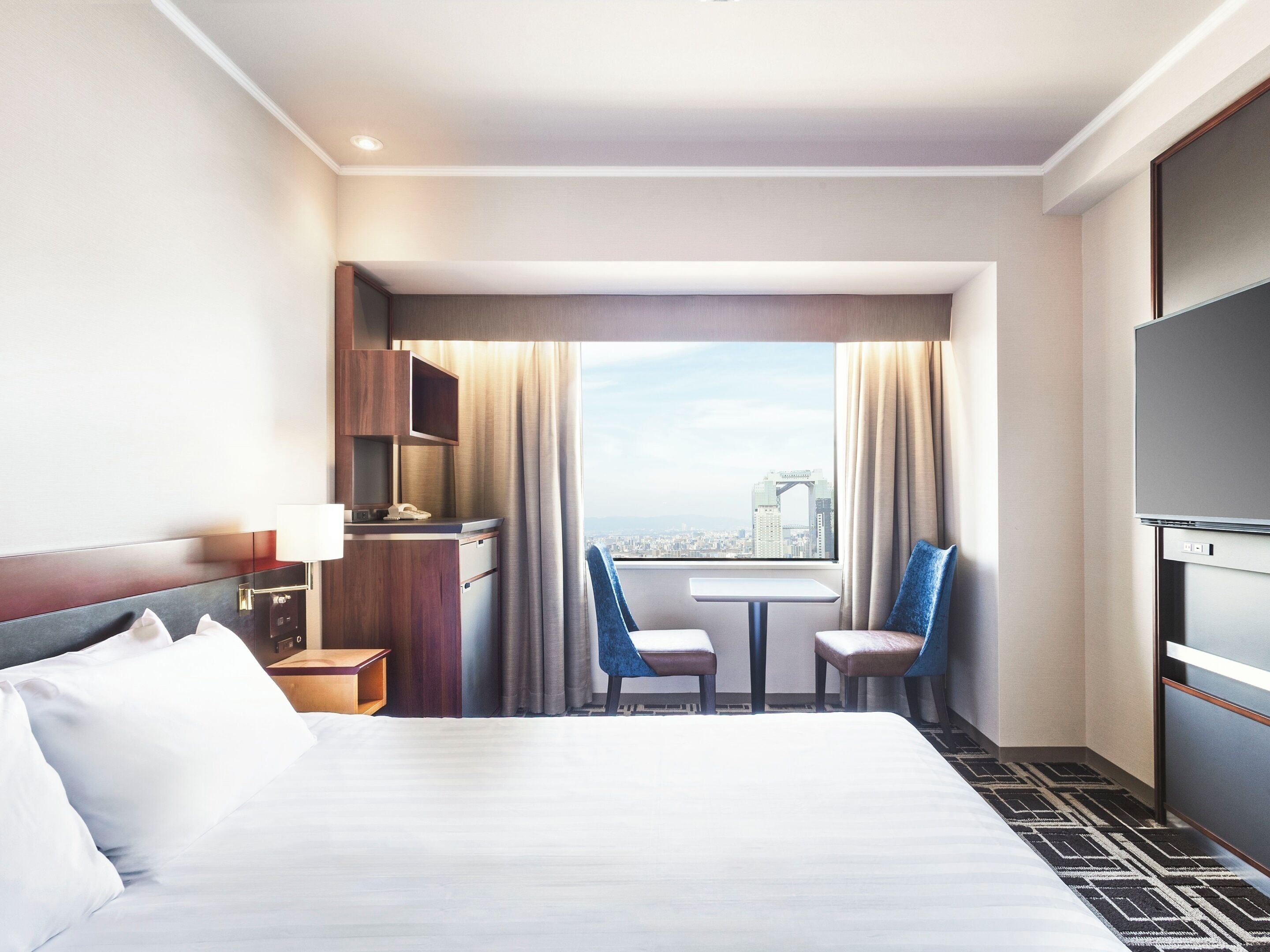 A hotel room with a bed, a desk, two chairs, a TV, and a window with a city view.