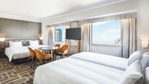 Habitación de hotel con dos camas, una mesa de comedor con sillas, un televisor y ventanas amplias con cortinas.