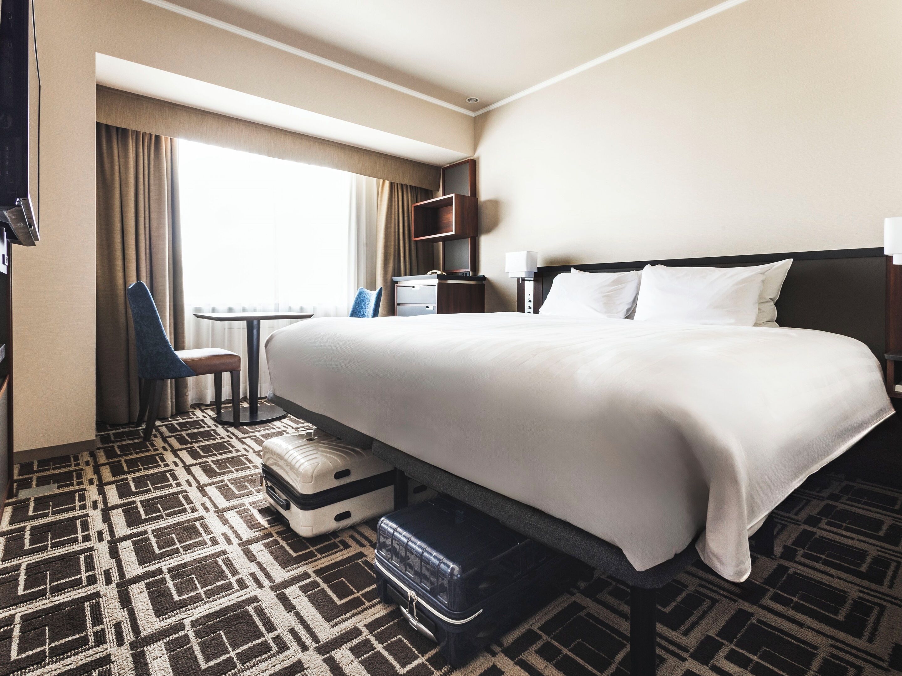 A hotel room with a large bed, a desk, a chair, a suitcase, and a window with curtains.