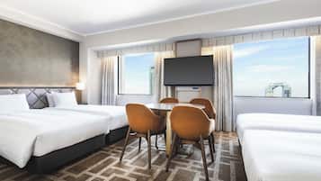 A hotel room with two beds, a dining table with chairs, a flat-screen TV, and large windows with a cityscape view.