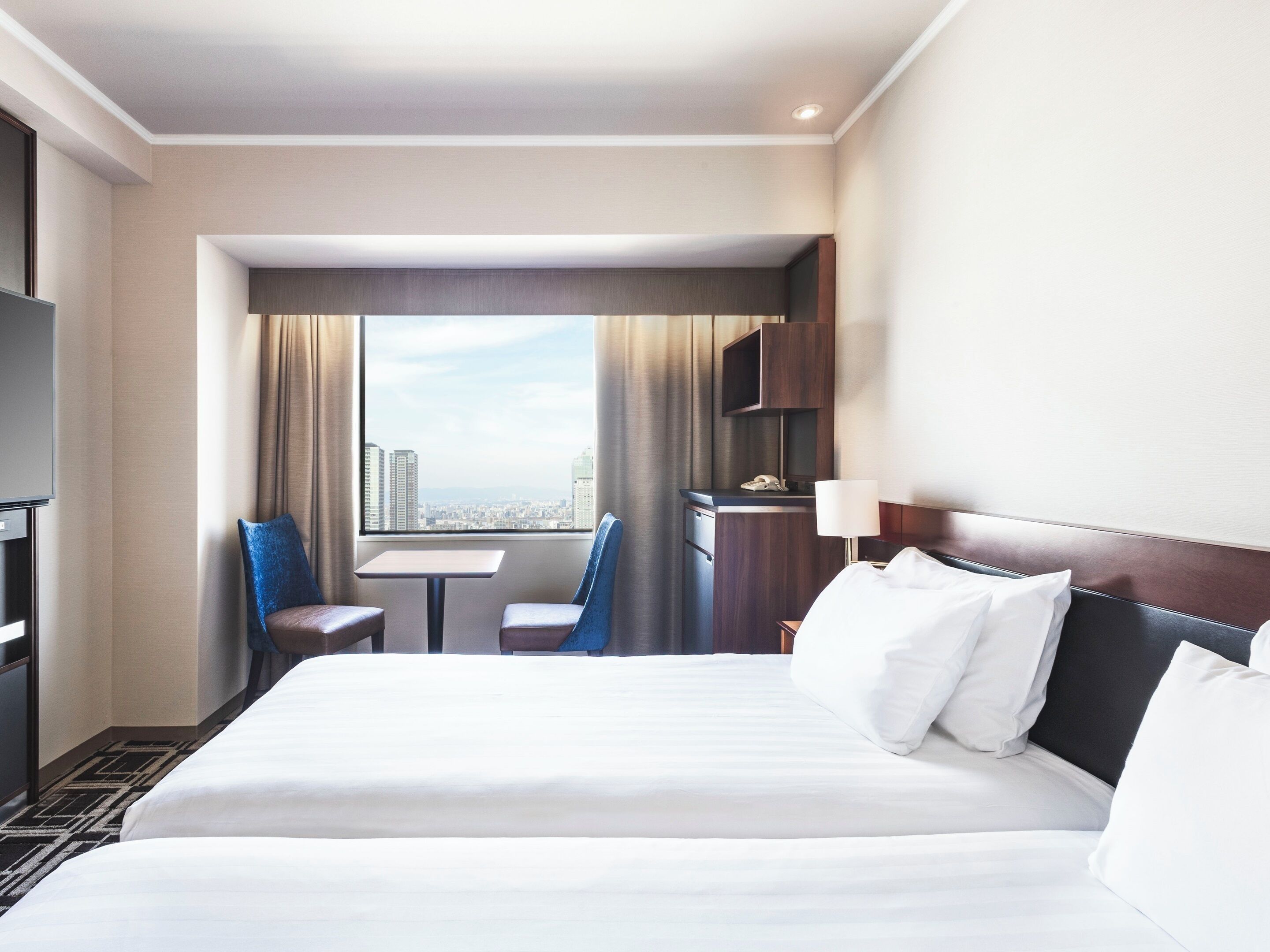 A hotel room with a large bed, a desk, two chairs, a TV, and a city view.