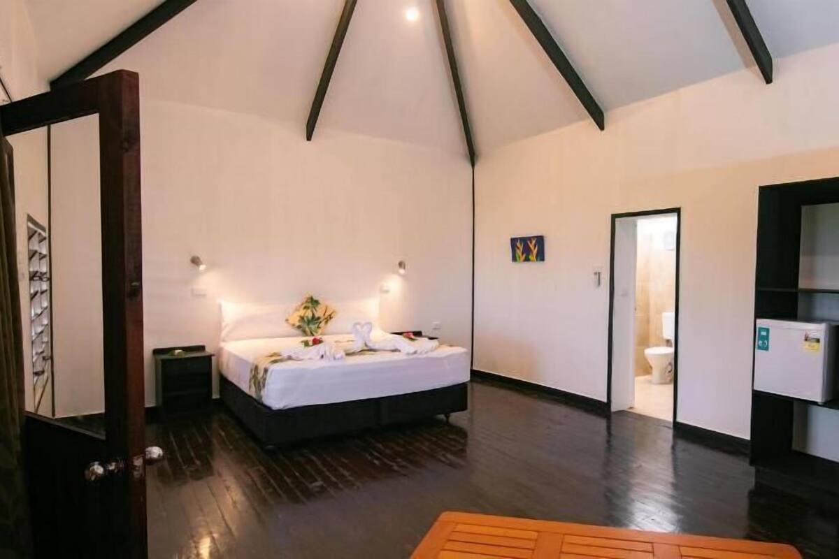 Una habitación amplia con cama, suelo de madera, una puerta que da a un baño y una mesita pequeña.