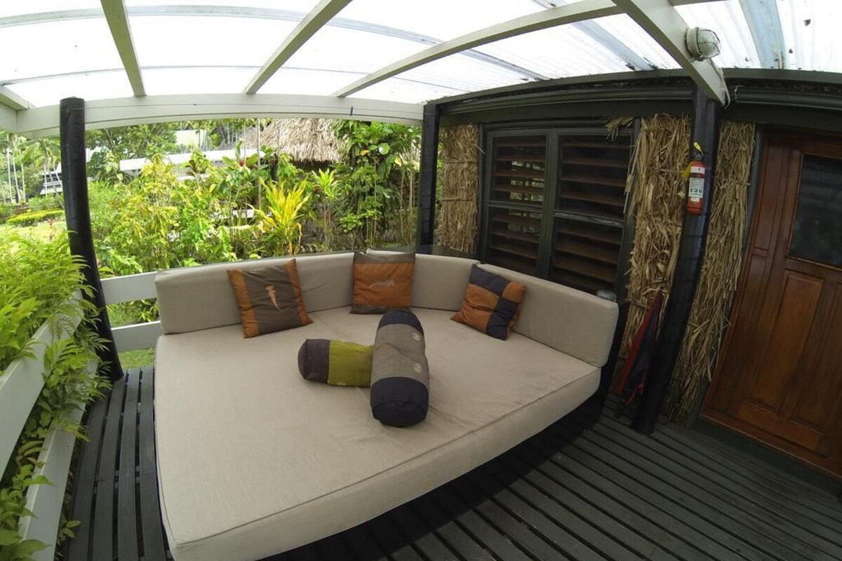 A curved sofa with multiple cushions in a covered outdoor area with a wooden deck and lush greenery visible through the transparent roof.