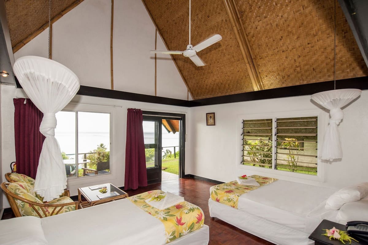 Una habitación con dos camas, ventilador de techo, ventana con persianas y vistas al exterior con vegetación.