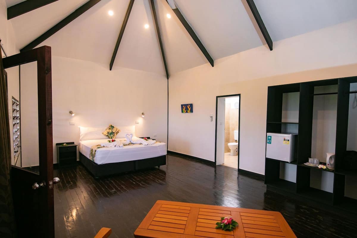 Un dormitorio con cama, mesitas de noche, suelo de madera, techo con vigas y una puerta que da al baño.