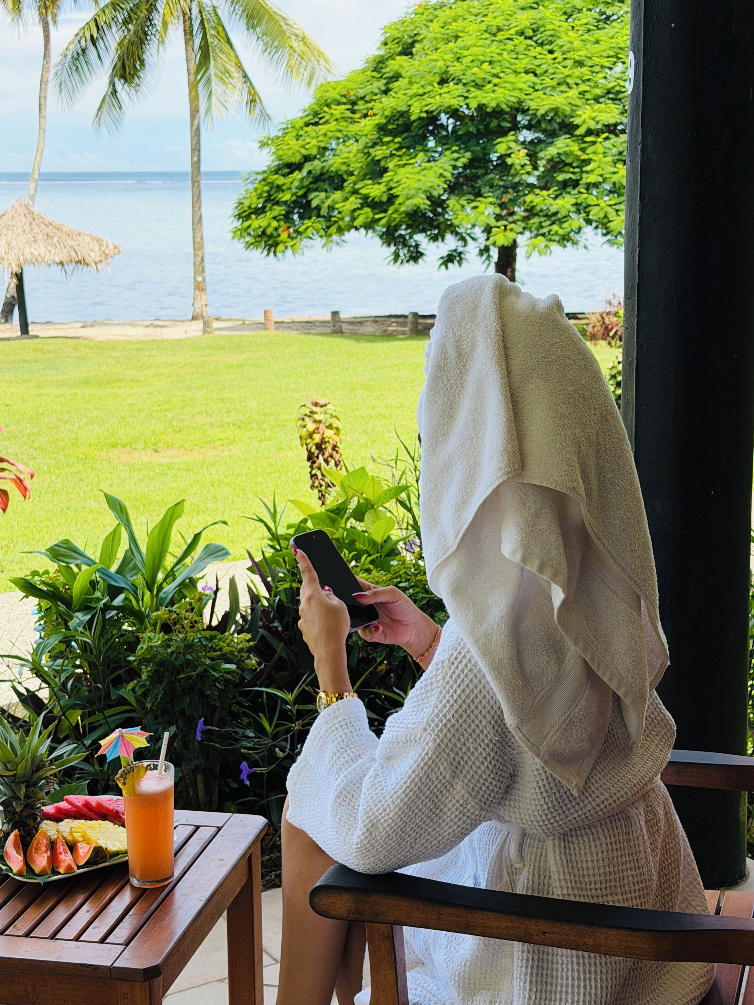A person using a smartphone, seated at a wooden table with a tropical drink and fruit, overlooking a grassy area with a palm tree and a view of the sea.
