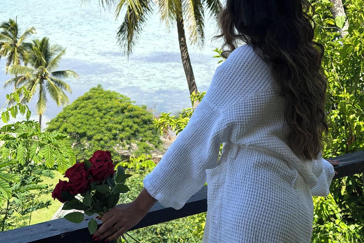 Una persona vestida de blanco, de pie junto a una barandilla, contempla una vista costera con palmeras y una extensión de agua.