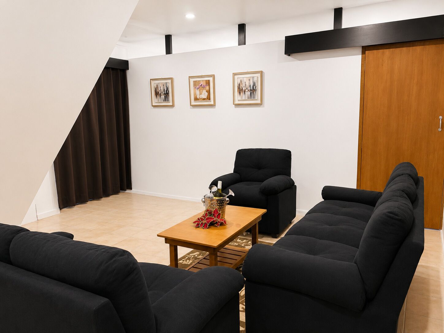 A modern living room with a black sofa set, a wooden coffee table, and a black armchair. There are three framed artworks on the wall, and a wooden door is visible.