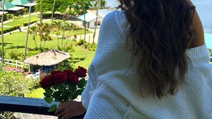 Una persona de cabello largo sostiene un ramo de rosas rojas, con vistas a un paisaje tropical con palmeras y el océano.