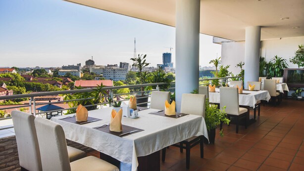 A rooftop restaurant with tables set for dining, overlooking a cityscape with buildings and greenery.