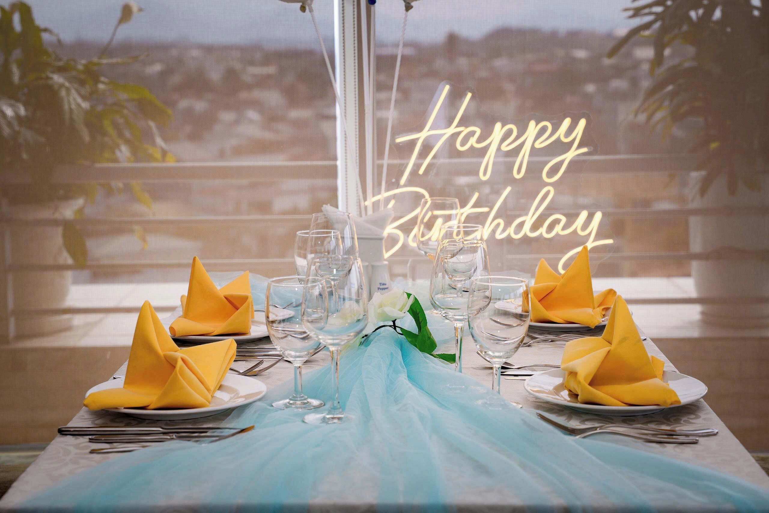 A table set for a birthday celebration with a neon sign, a view of a cityscape, and a blue tablecloth.