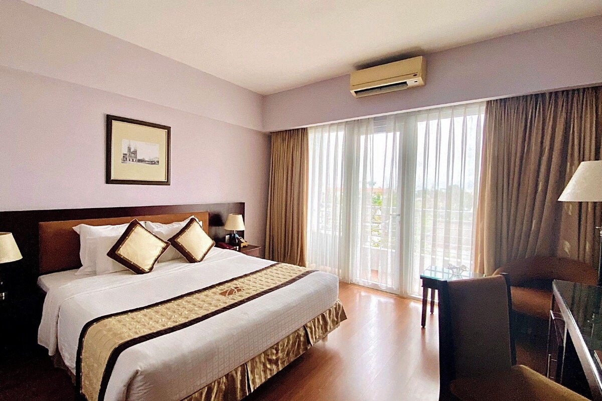A hotel room with a large bed, two bedside tables with lamps, a framed picture on the wall, and a view of the outdoors through the window.