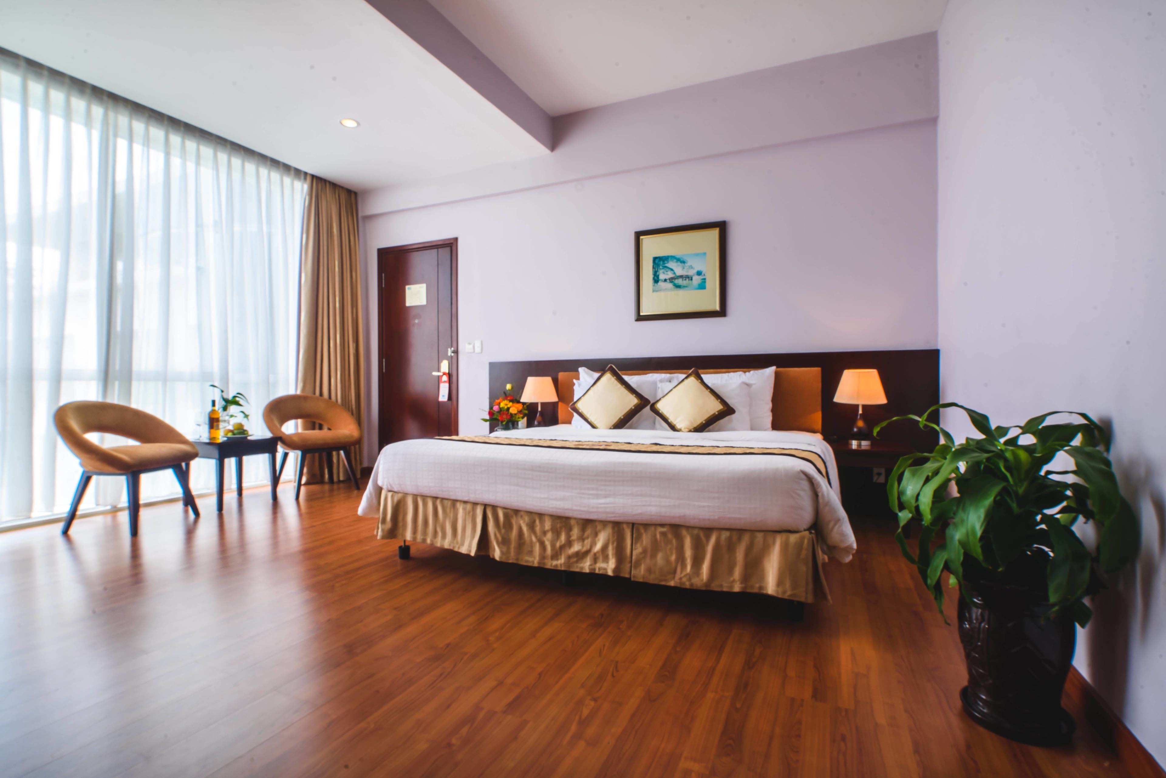Een hotelkamer met een groot bed, twee stoelen, een tafel, een potplant en een schilderij aan de muur.