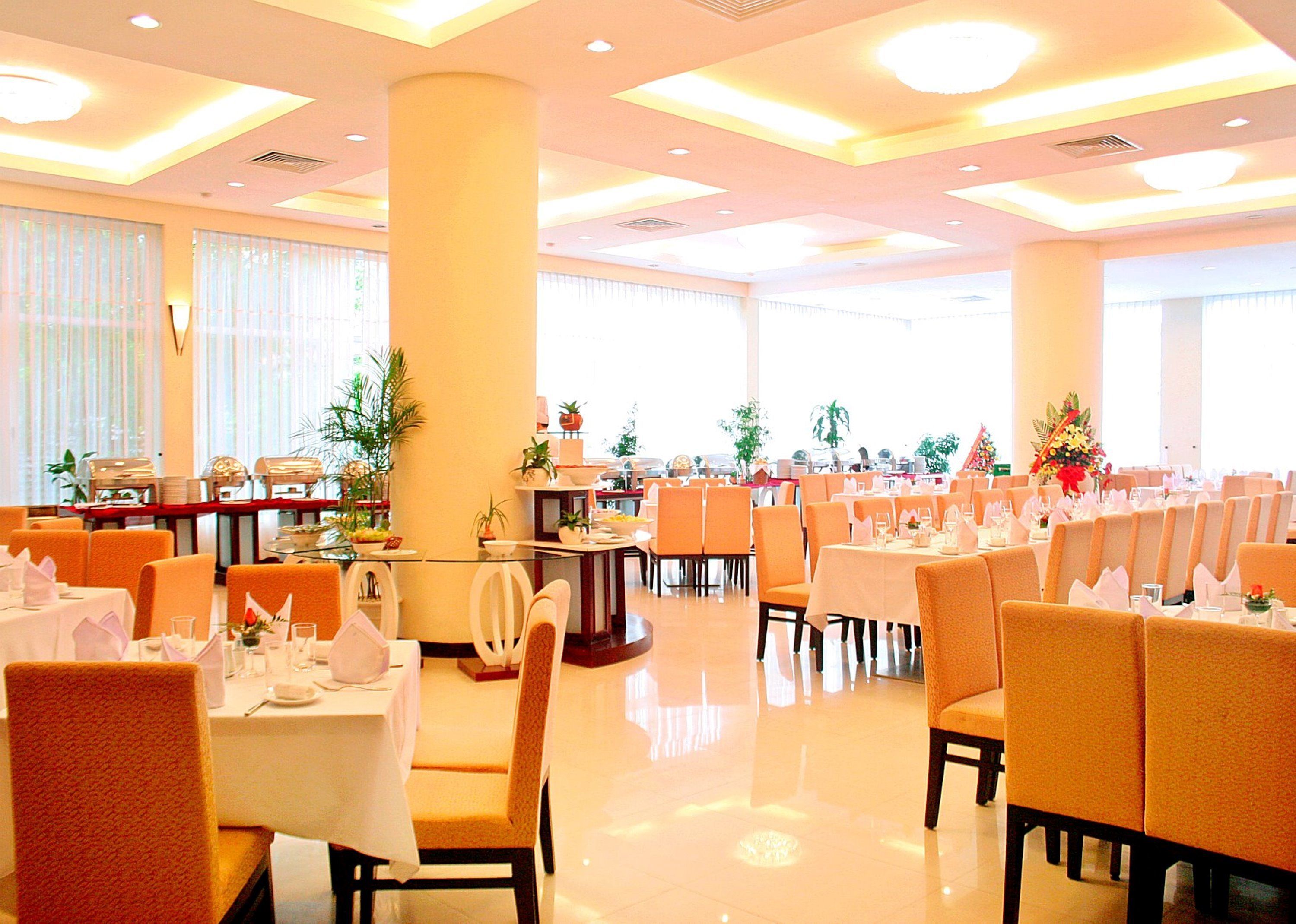 A spacious dining area with multiple tables and chairs, large windows, and decorative plants.
