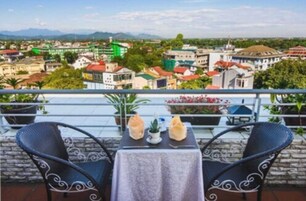 Un balcón con dos sillas y una mesa, con vista a un paisaje urbano con edificios y áreas verdes.
