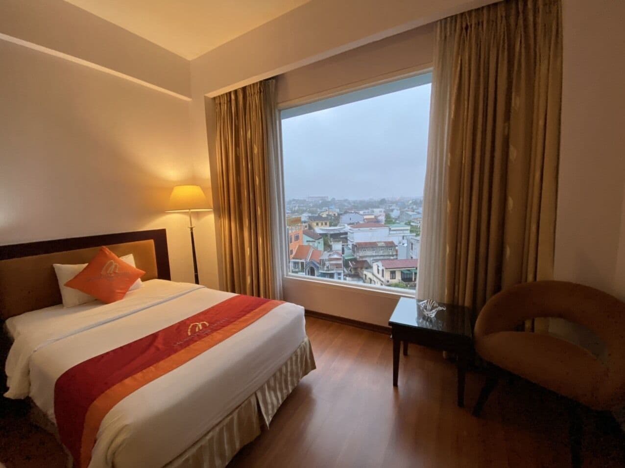 A hotel room with a large bed, a view of the city, and a chair by the window.