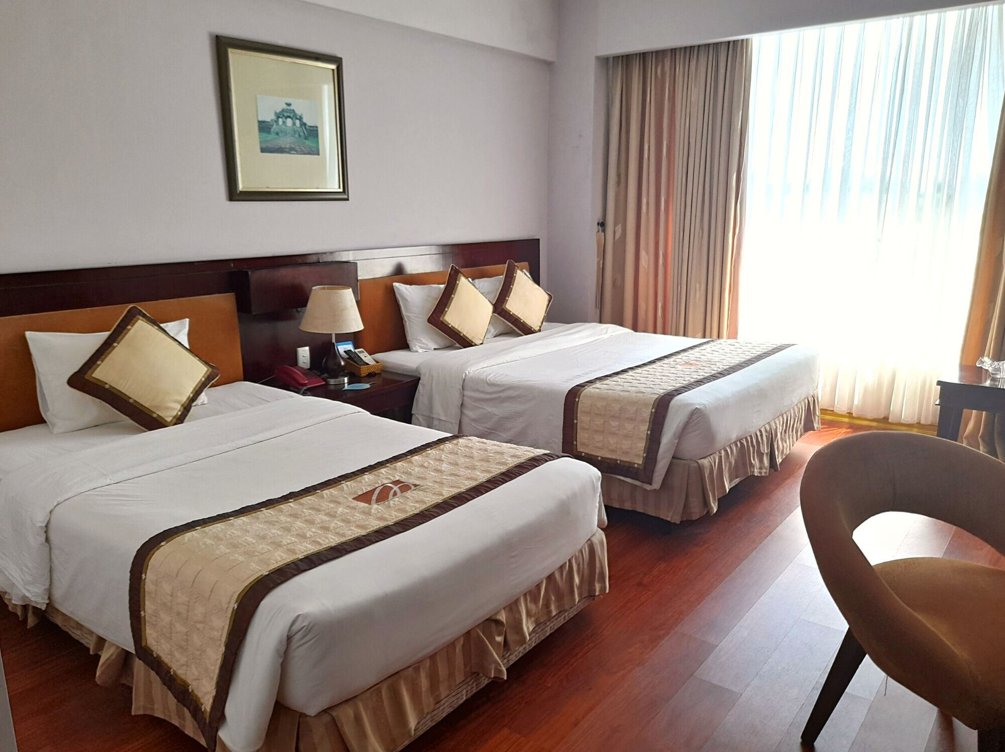 Habitación de hotel con dos camas, una mesita de noche con lámpara, una silla y un ventanal con cortinas.