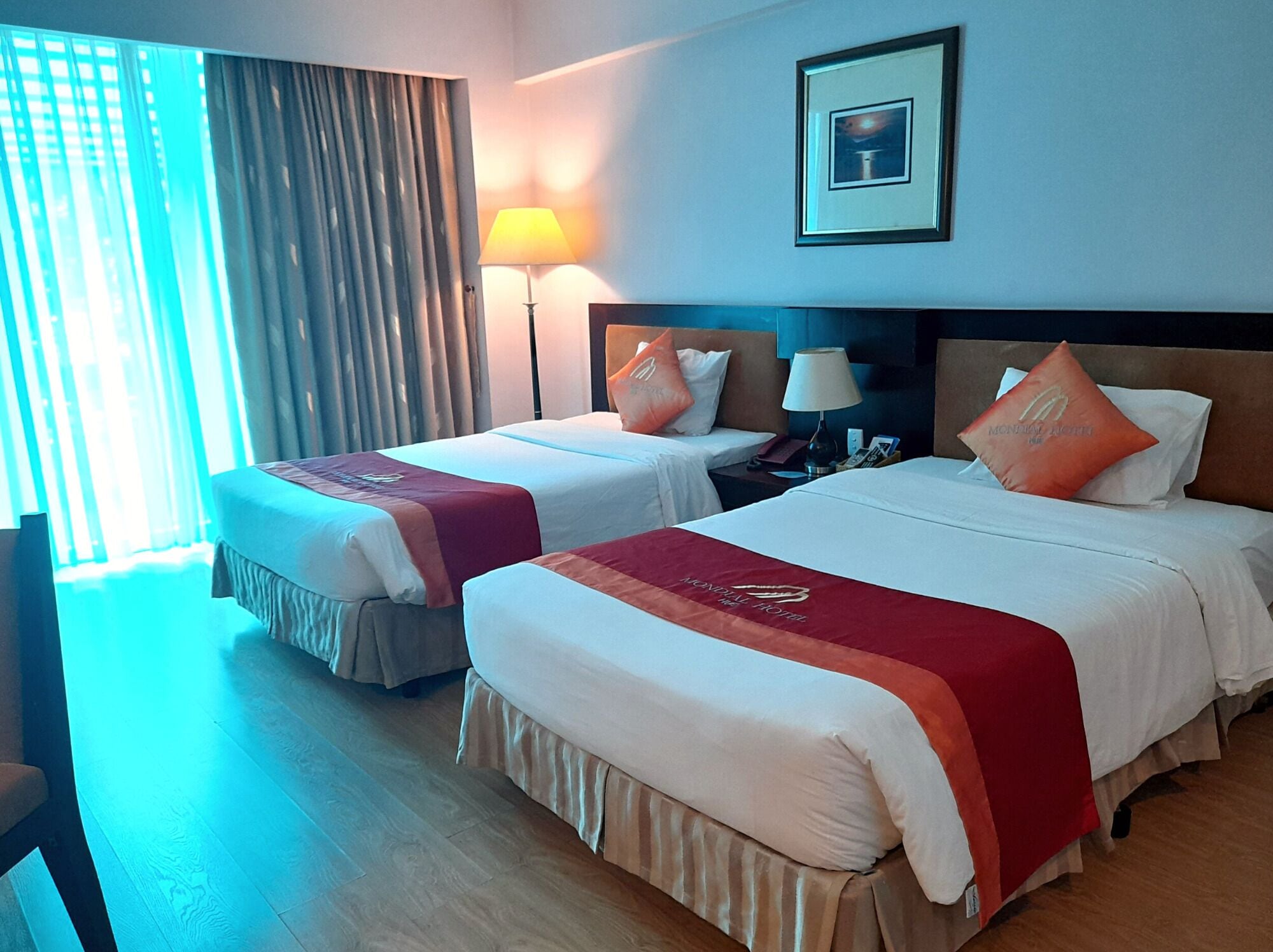 Habitación de hotel con dos camas, una mesita de noche, una lámpara, un cuadro en la pared y una ventana con cortinas.