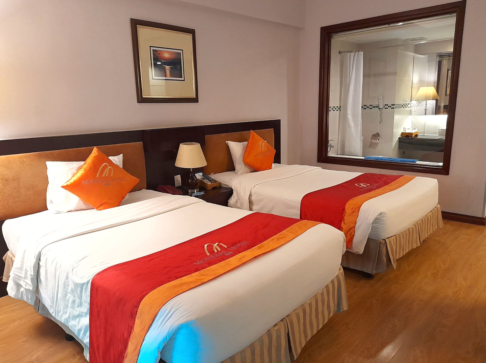 A hotel room with two beds, a nightstand with a lamp, a framed picture on the wall, and a large mirror.