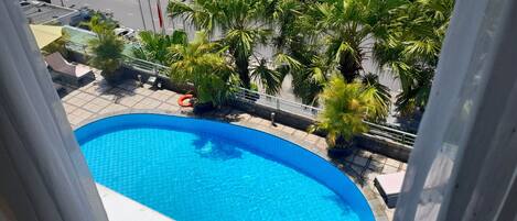Vista de una piscina rodeada de palmeras y una calle con edificios al fondo.