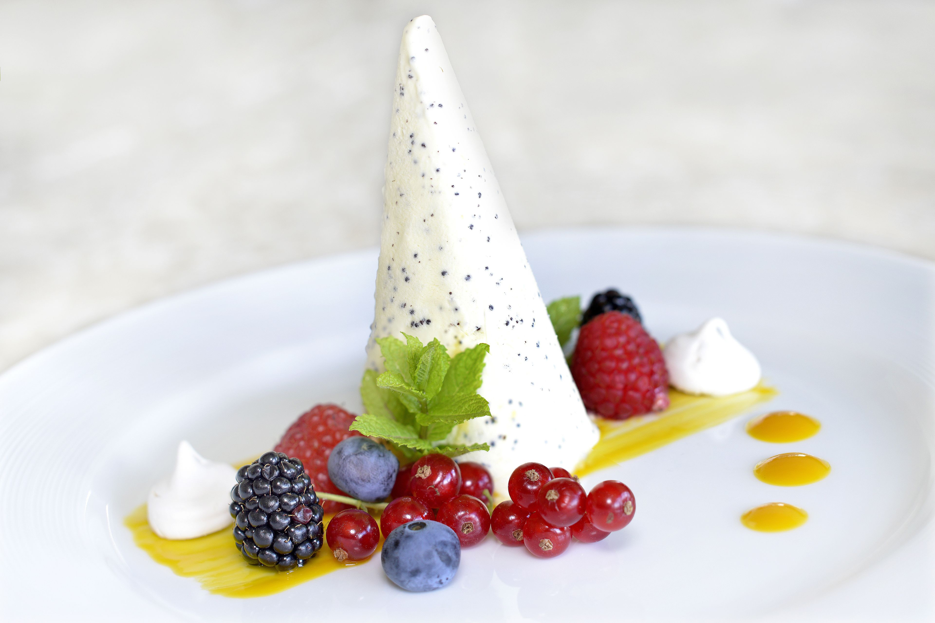 A dessert with a cone-shaped ice cream topped with berries and a mint leaf, served on a white plate with a drizzle of syrup.