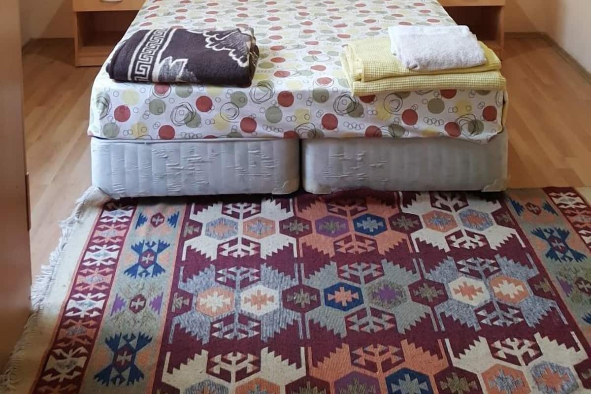 A bedroom with a bed, a nightstand, a kettle, and a patterned rug.