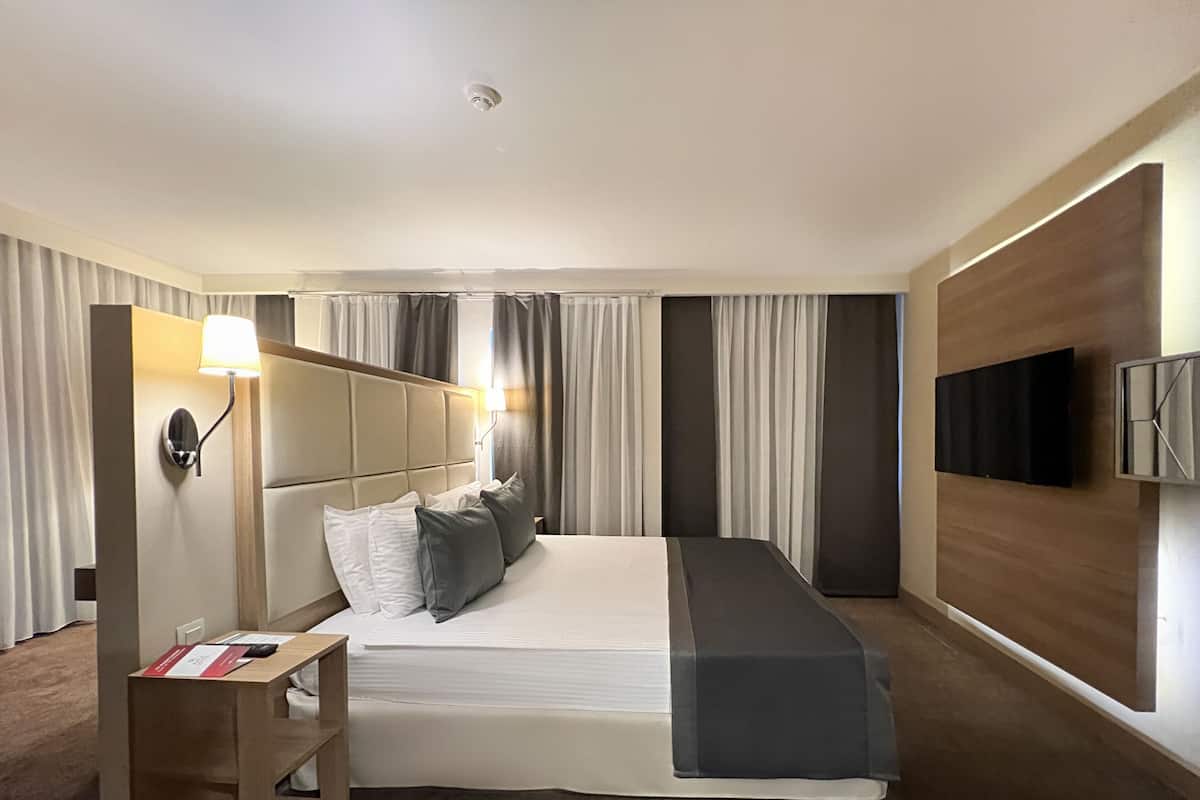 A hotel room with a large bed, a bedside table with a lamp, a TV mounted on the wall, and curtains on the windows.