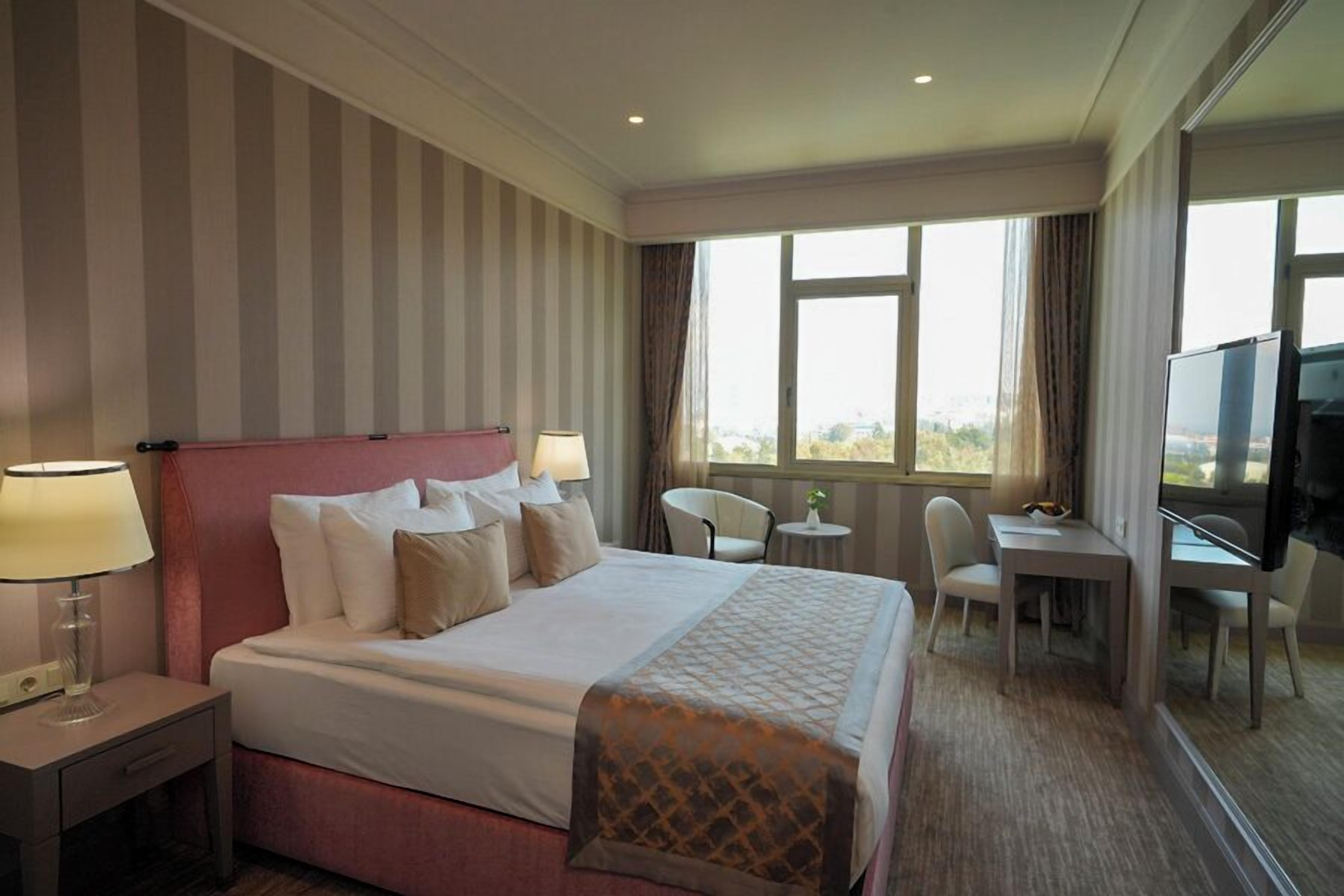 A hotel room with a large bed, bedside tables with lamps, a desk with a chair, a TV, and a view of the city through large windows.