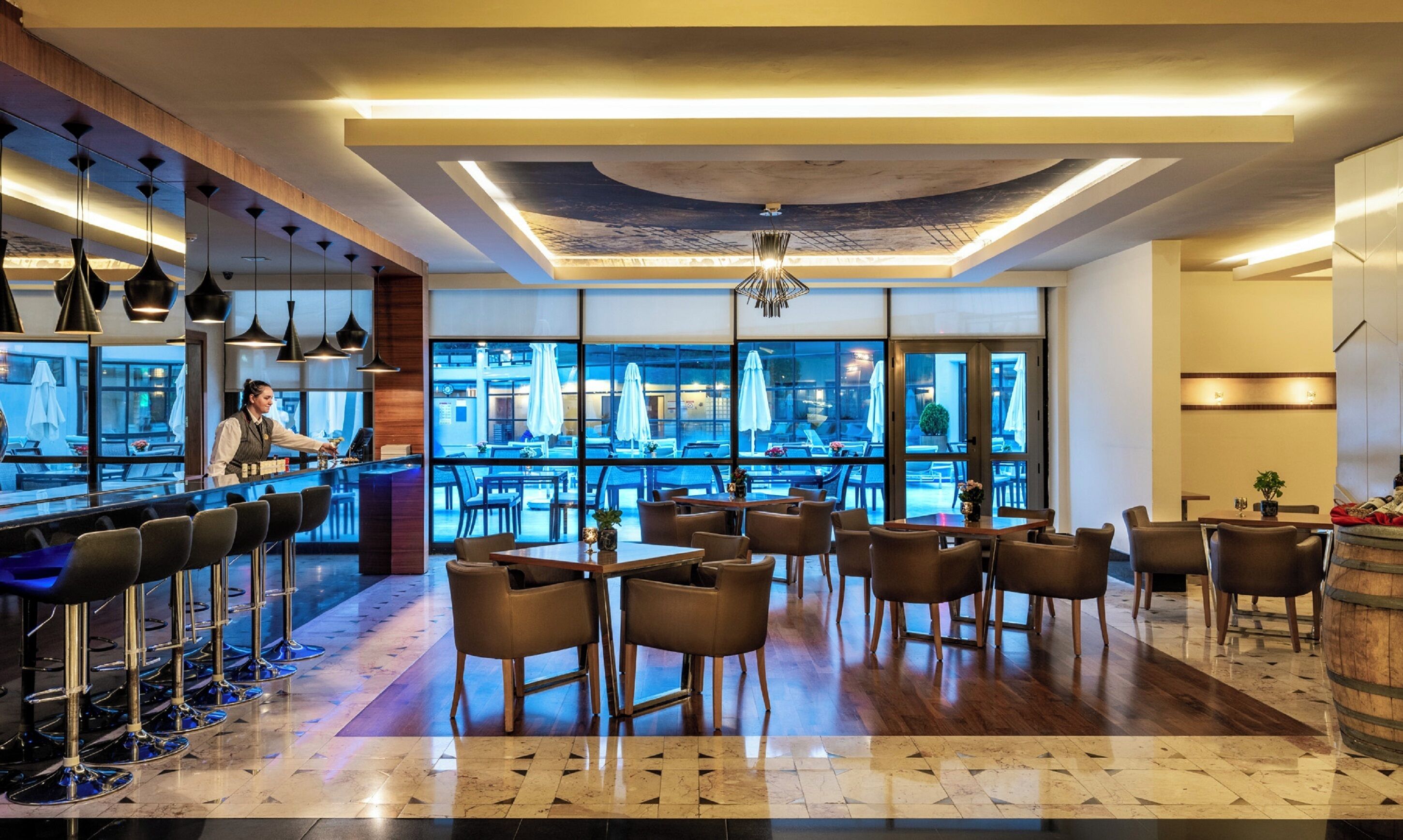 A modern bar area with a bartender, a seating area with tables and chairs, and large windows offering a view of the outdoor seating area.