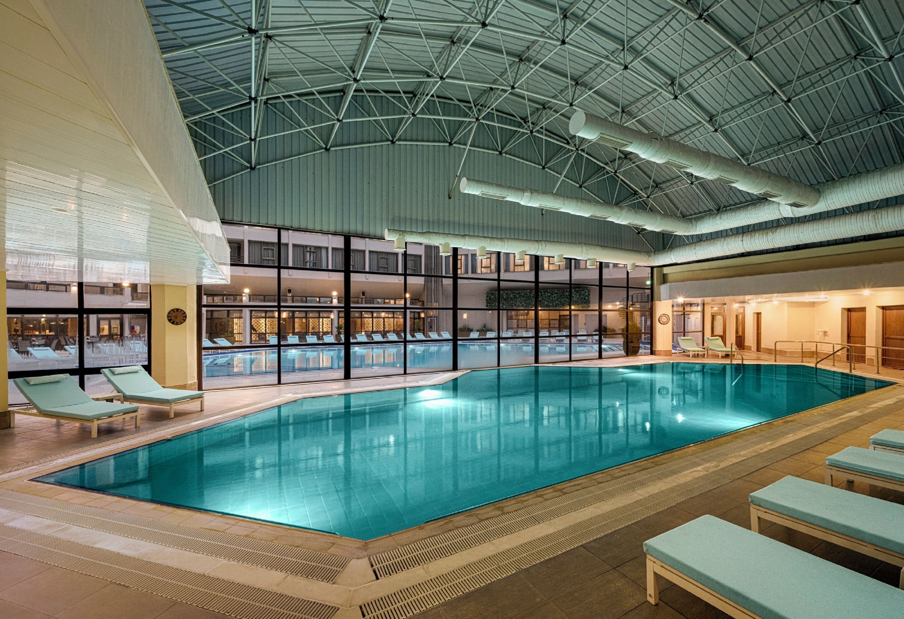 An indoor swimming pool with lounge chairs, a high ceiling with visible structural beams, and a view of the outside through large windows.