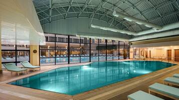 An indoor swimming pool with lounge chairs, a high ceiling with visible structural beams, and a view of the outside through large windows.
