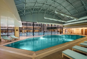 Piscina interna com espreguiçadeiras, teto alto com vigas aparentes e vista para o exterior através de grandes janelas.